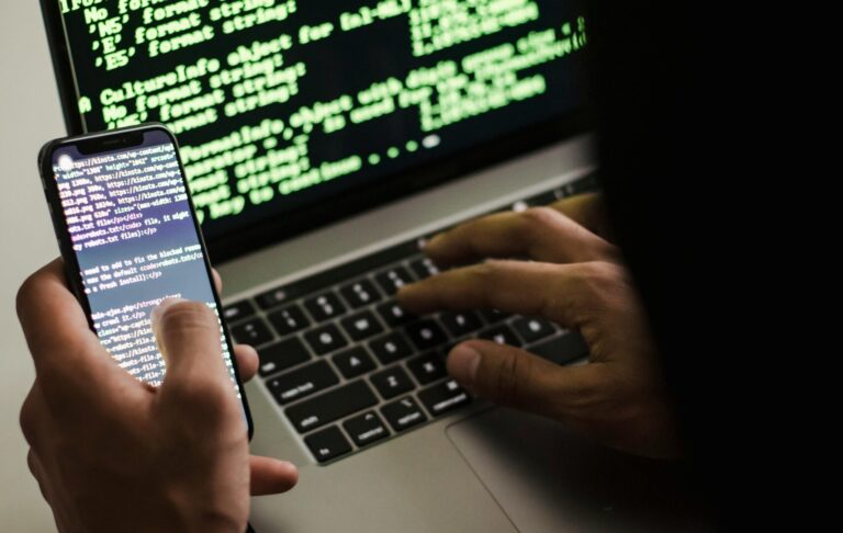 A hooded figure engaged in hacking using a laptop and smartphone in low light.